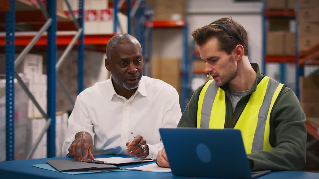 Manager With Male Worker Wearing Headset Sitting At Desk Working On Laptop In Busy Warehouse