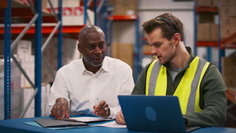 manager with male worker wearing headset sitting at desk working on laptop in busy warehouse