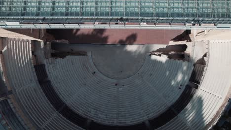 Toma-Superior-De-Un-Dron-Del-Antiguo-Teatro-Naranja,-La-Simetría-De-La-Toma-Es-Magnífica,-Vemos-El-Piso-De-Madera,-Las-Gradas-De-Piedra-Y-El-Techo-De-Vidrio.