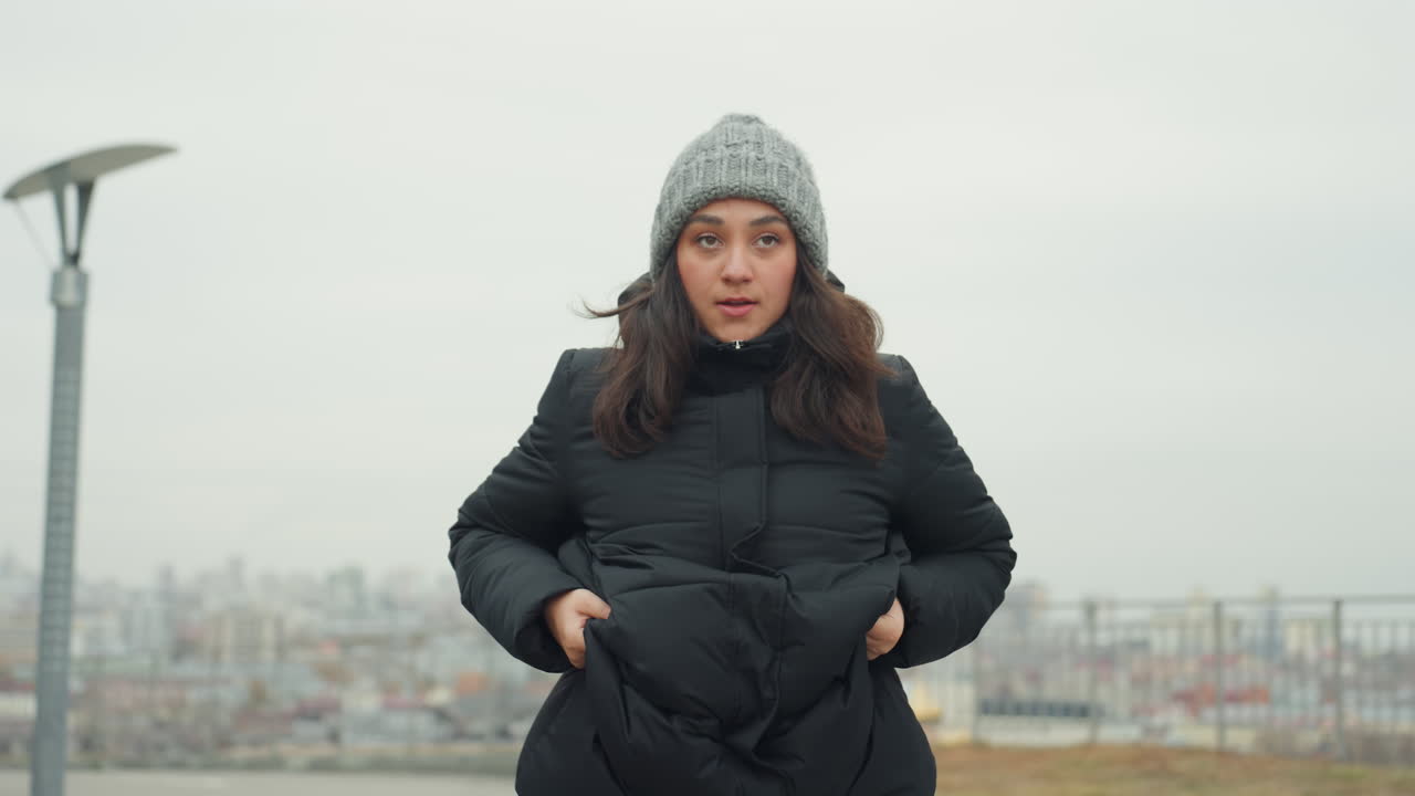 Primo piano di donna bianca che balla con espressione, gesti facciali intensi e movimenti delle mani, sollevamento della gamba e prova concentrata su una collina ventosa, dettagli del cappotto invernale e del berretto, skyline urbano cupo, riflessivo