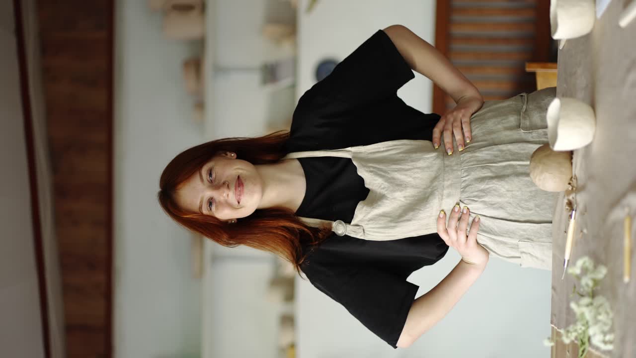 una chica alfarera de cabello rojo en y delantal mira hacia adelante, sus manos están colocadas en su cintura