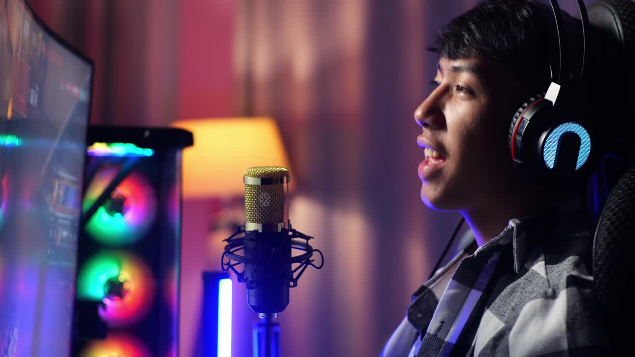 vista lateral de cerca de un niño asiático que juega a un juego a través de la red en una computadora personal, hablando con los participantes en el micrófono. videojuego de transmisión en vivo, escritorio iluminado por luz de tira de led rgb