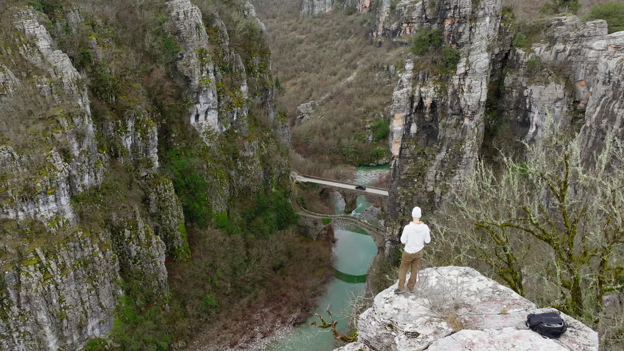 Una persona è in piedi su una scogliera e guarda giù verso l'antico ponte in pietra di Kokkorou sul fiume turchese in Epiro, Grecia.