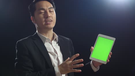 블랙 스크린 스튜디오에서 말하는 동안 녹색 스크린 태블릿을 들고 가리키는 비즈니스 슈트를 입은 아시아 스피커의 클로즈업