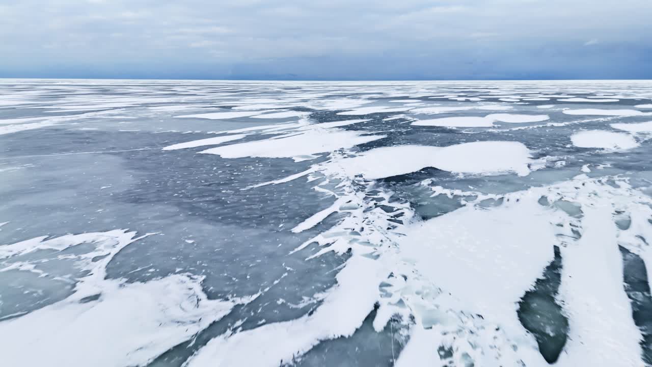 Vista aerea di lastre di ghiaccio innevate e scuri motivi di congelamento che si estendono verso un orizzonte nuvoloso, formando una texture invernale astratta su un vasto paesaggio marino freddo.