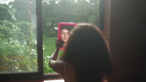 woman with curly hair holding pink mirror, reflecting her face by a window with green view outside