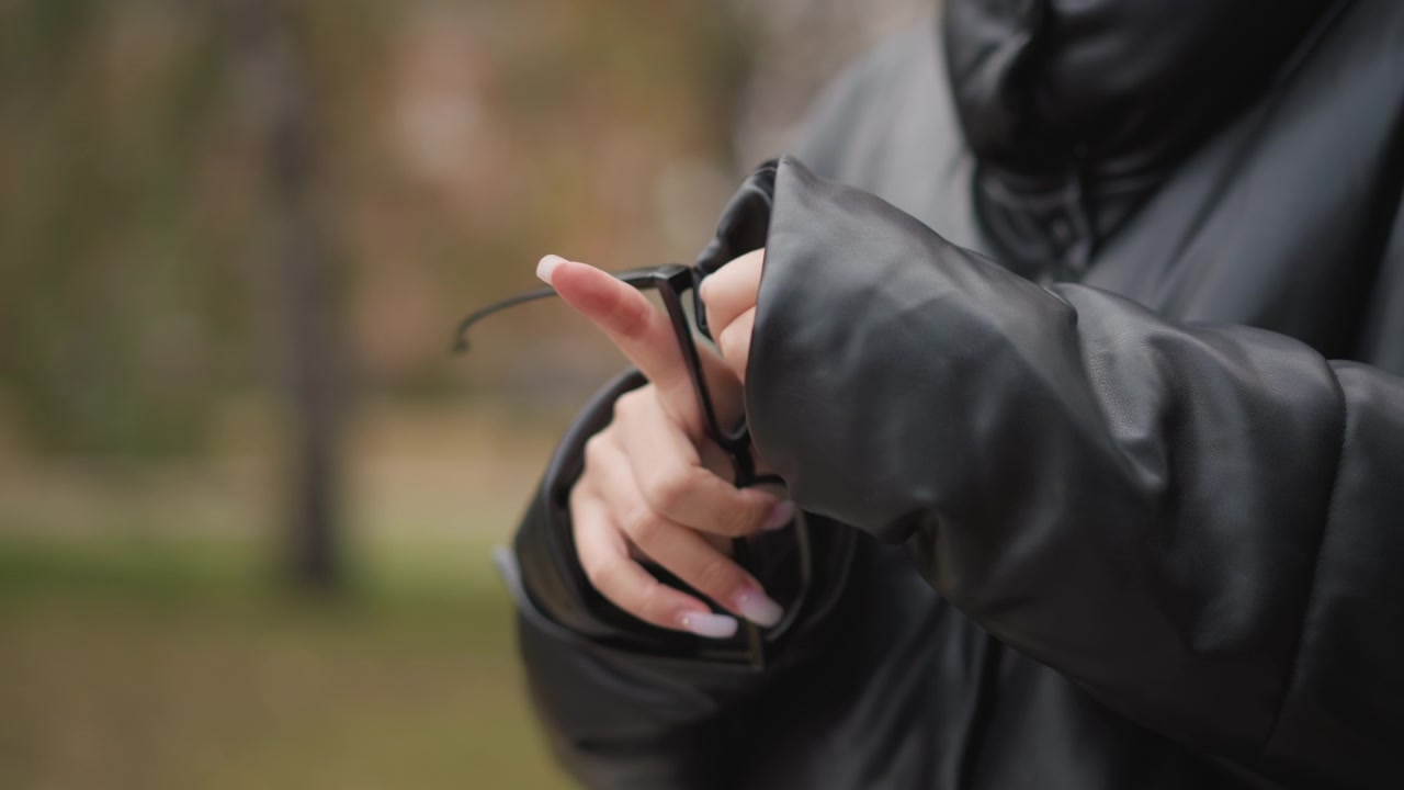 Close up of person wearing black leather jacket adjusting eyeglasses in hand. person manipulates glasses revealing their texture and form against outdoor park background with soft focus