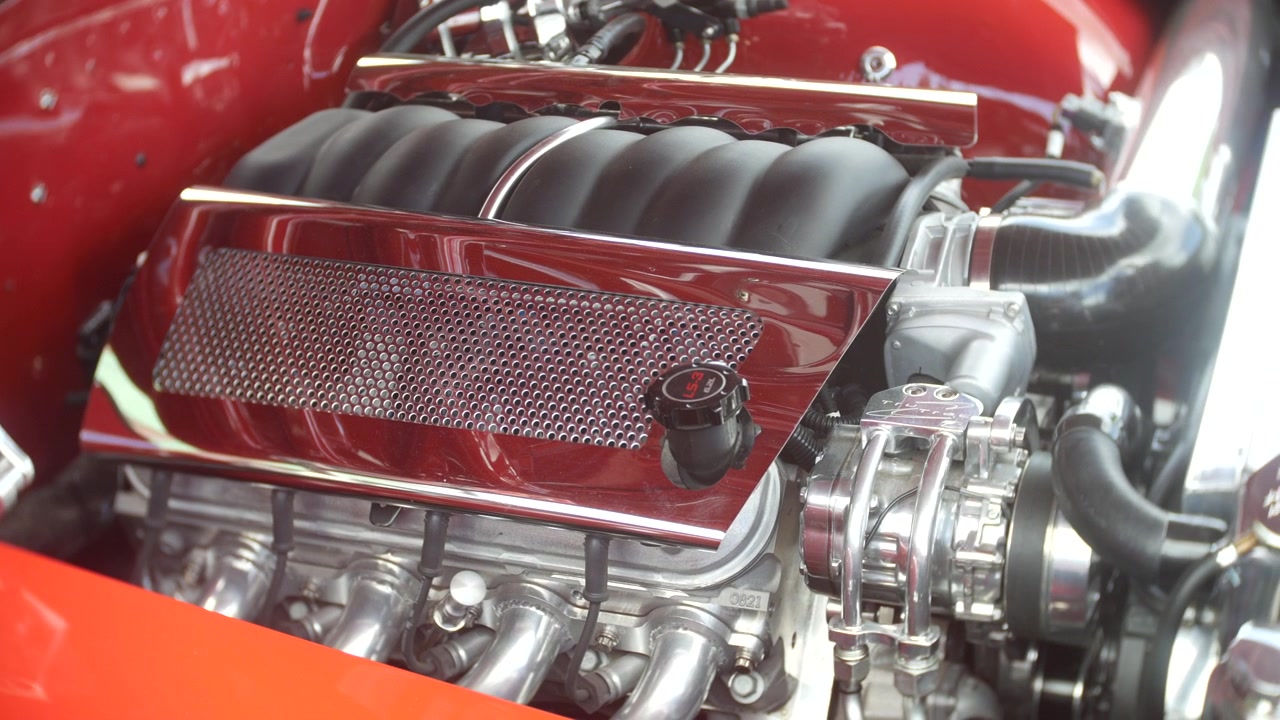 Motore a vista all'interno di una hot rod personalizzata mostra componenti LS3 lucidati e dettagli dell'aspirazione. La vista ravvicinata evidenzia tubi, cinghie, copertura in rete metallica e carrozzeria arancione.