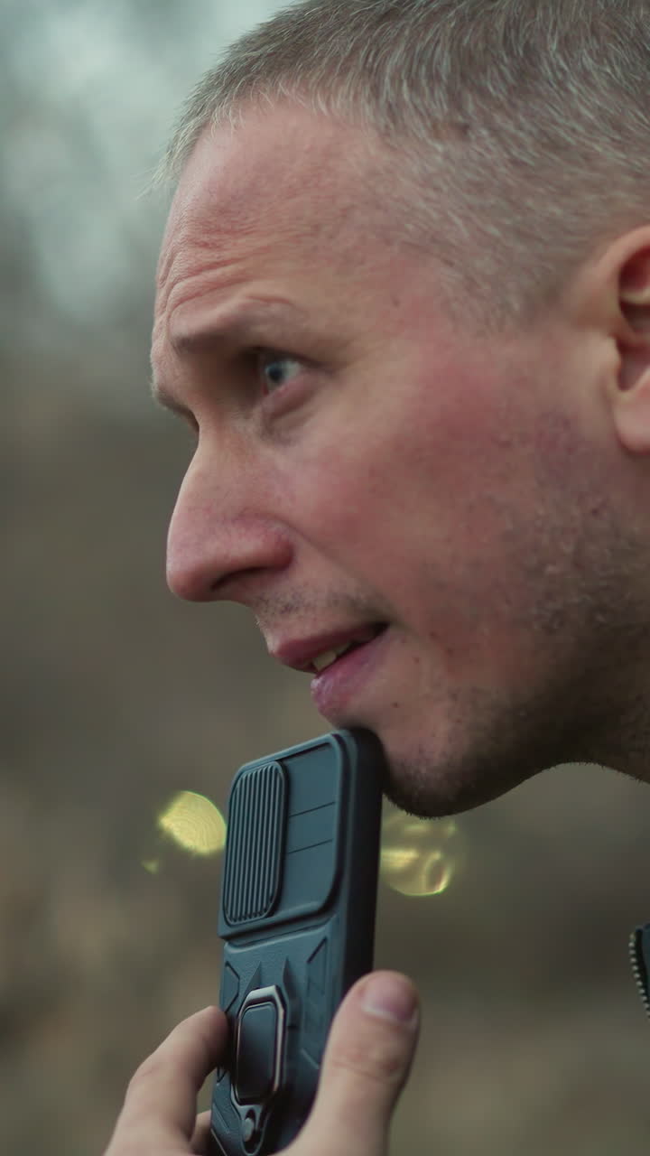 A close view of a man looking intensely to the side, holding a black smartphone near his mouth, the background features a forest, with two blur ball like yellow light seen