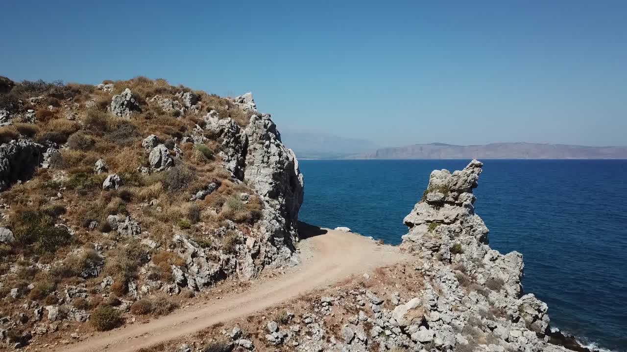 Dusty path winds through rocky cliff above deep blue sea with clear sky and distant land. Rugged terrain and natural formations create dramatic coastal scene in peaceful Santorini location