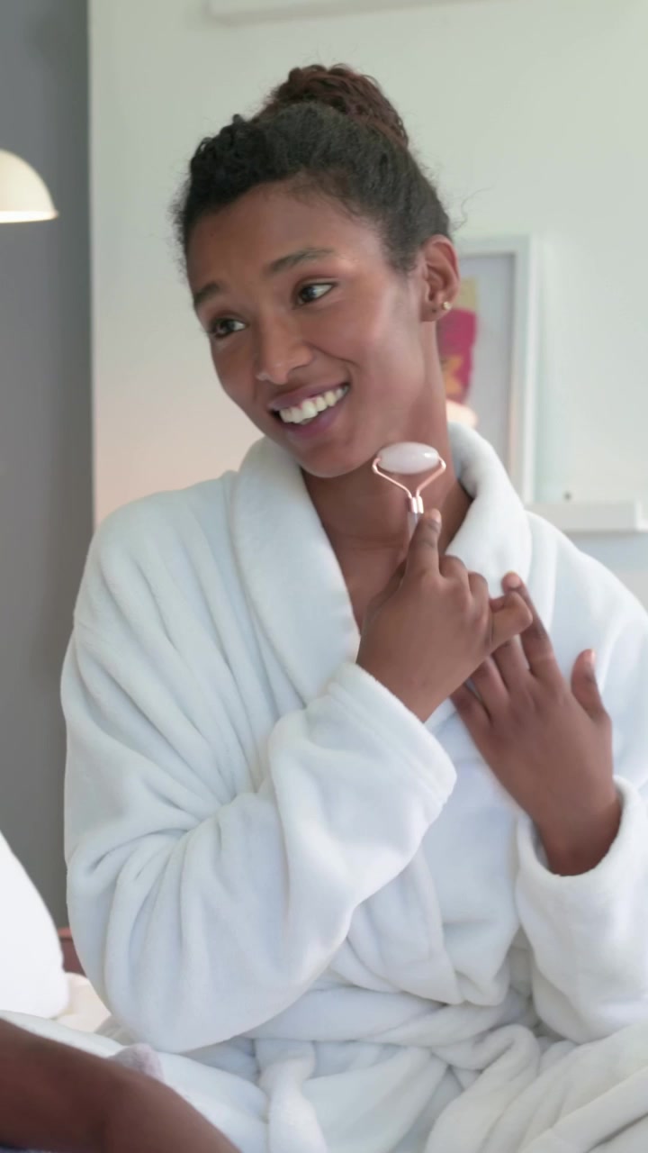 Vertical video: Talking offscreen, woman in robe using rose roller on jaw or neck for skincare at ho