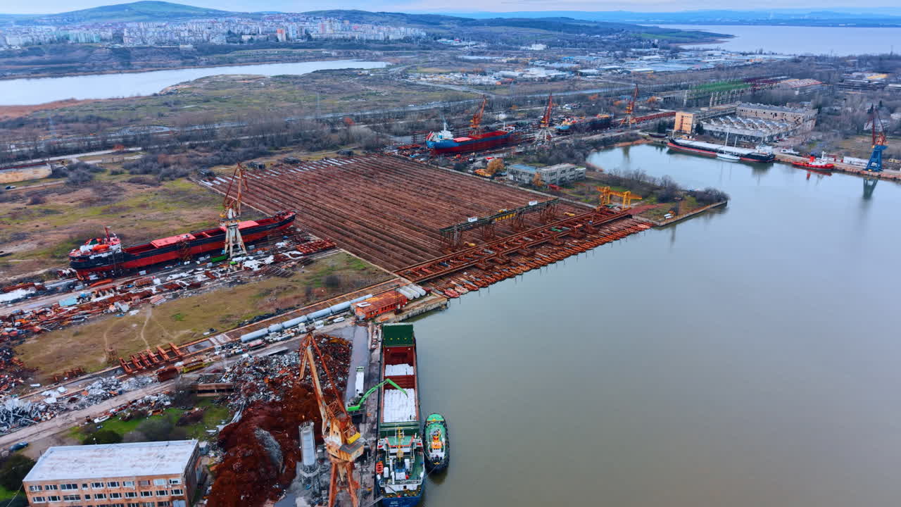 Paesaggio di cantiere navale industriale con nave da carico e depositi di metalli. Veduta aerea di un cantiere navale che presenta aree di riparazione navi, strutture metalliche e una nave da carico in fase di caricamento.