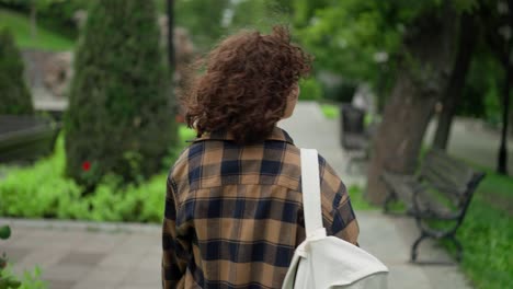 Rear-close-up-view-of-a-girl-with-curly-hair-in-a-plaid-shirt-walking-through-the-park-and-turning-back-during-her-walk-after-her-student-work