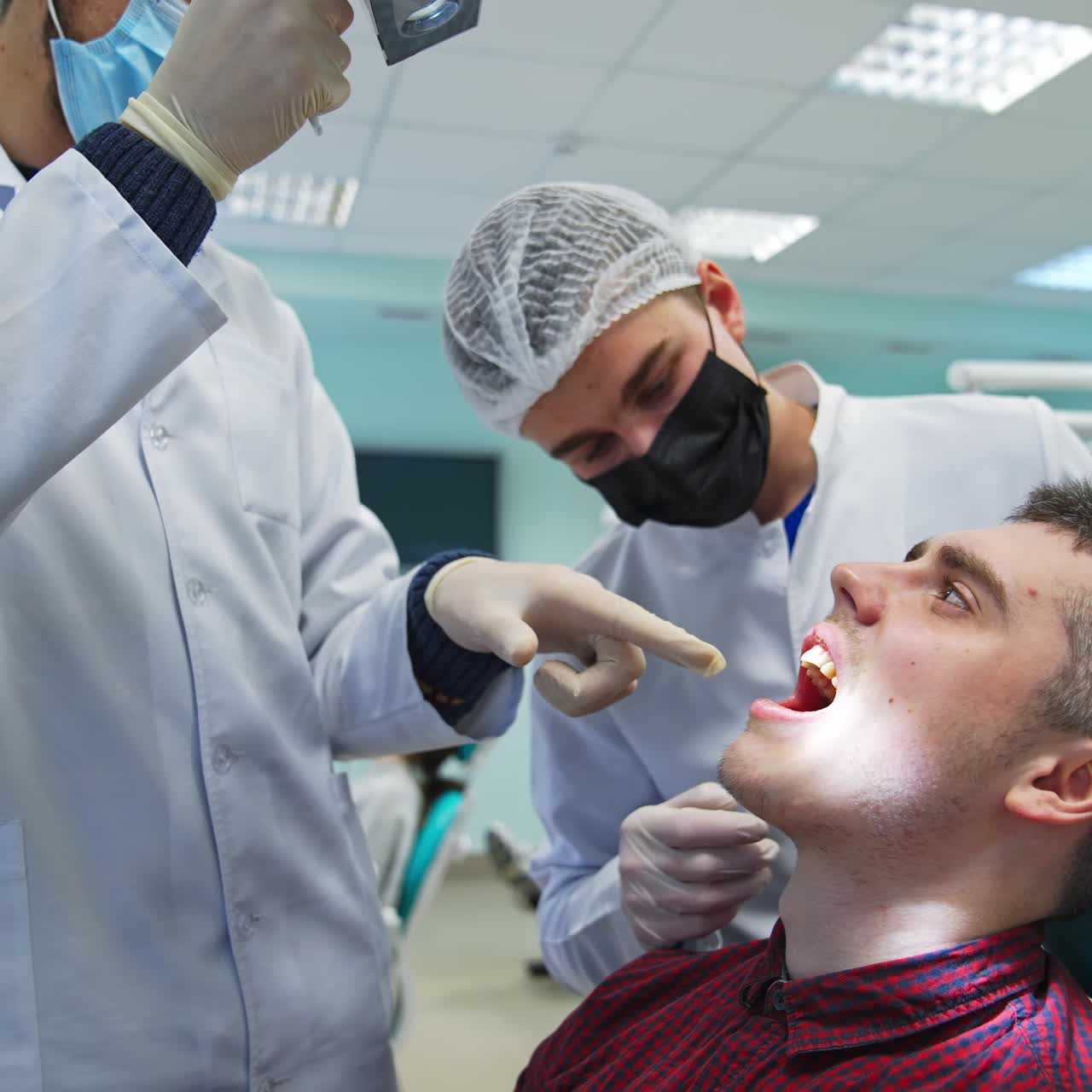 Male patient sitting in dentist chair with his mouth open. Two students bent over him, point at something in his mouth and comment. Classroom background