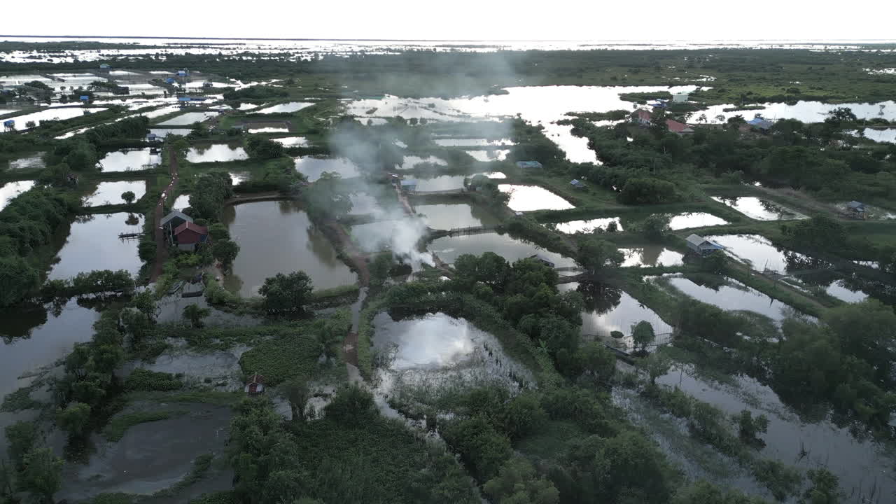 Aerial view of Tonlé Sap wetlands with smoke rising from a small homestead among reflective ponds
