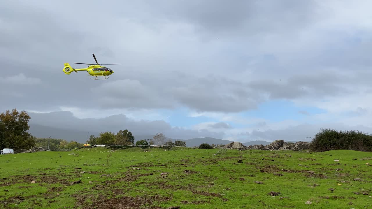 filming in slow motion in a rural area the takeoff of a yellow emergency helicopter, we see how it is already ascending and the sky is cloudy, in the tree area there are lights from an ambulance