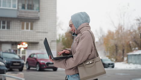 side view of light skin lady dressed in warm hooded jacket holding laptop and simultaneously speaking on phone while standing outside on snowy street with cars and buildings in background