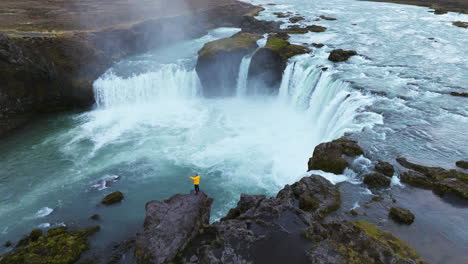 Vista-Aérea-De-Un-Hombre-Al-Borde-De-Un-Acantilado-Cerca-De-La-Cascada-Godafoss-En-Islandia