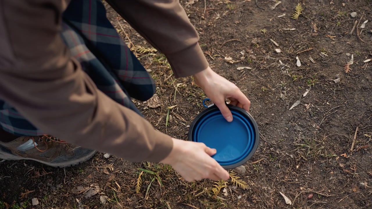 Persona che indossa scarpe da esterno e pantaloni a quadri si inginocchia su terreno sterrato, maneggiando una ciotola pieghevole blu aperta. La scena mostra dettagli di mani, piedi, terreno con piccole piante e ramoscelli.