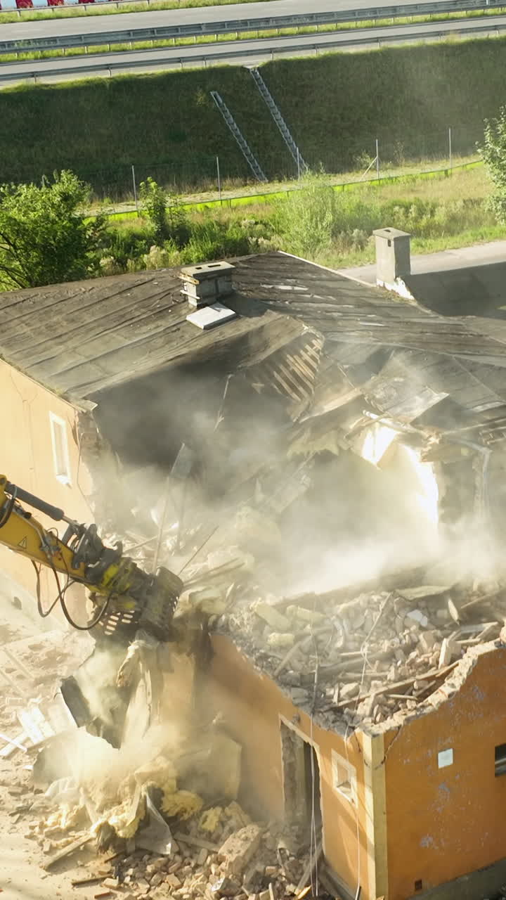 Une puissante pelleteuse jaune utilise un concasseur hydraulique pour démolir un ancien bâtiment, créant de la poussière et des débris. Plan cinématographique en plongée du processus de rénovation urbaine et de démolition.