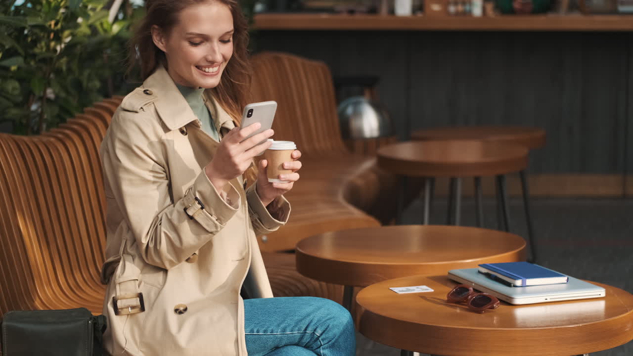 Caucasian female student using smartphone and drinking coffee outdoors.