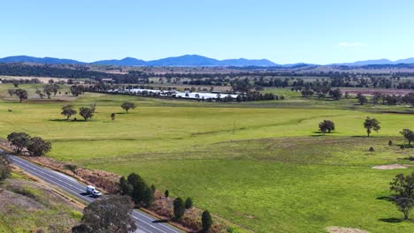 drone camera smoothly pans above a country road, revealing expansive green fields, scattered trees, distant hills, and clear blue skies in rural new south wales