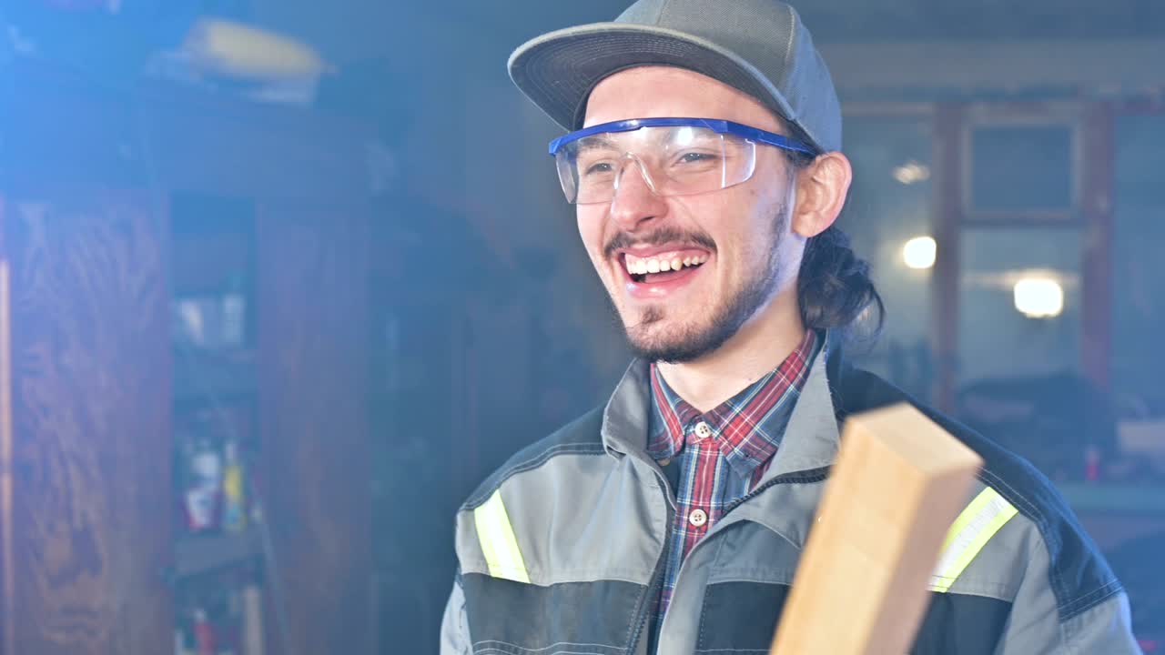 joven carpintero barbudo concentrado con gafas de protección y una gorra comprueba la pieza de trabajo