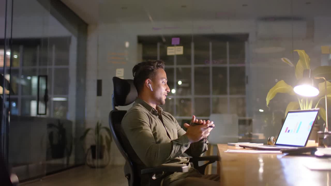 Tired happy biracial businessman listening to music with earphones in office at night, slow motion