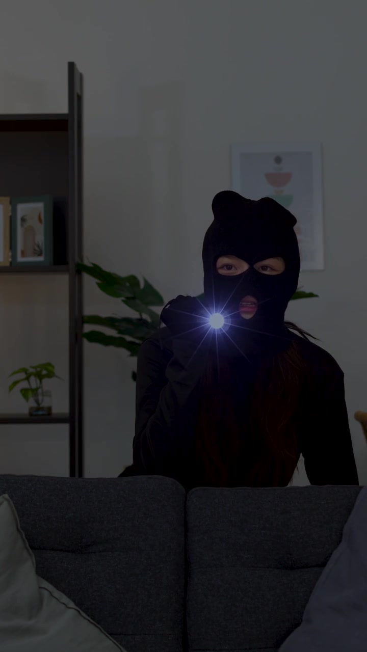 A masked intruder cautiously scans a dark living room using a flashlight during a burglary