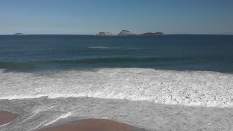 Seitliche-Luftaufnahme-Von-Wellen,-Die-Am-Strand-Von-Ipanema-In-Rio-De-Janeiro-Mit-Inseln-Direkt-Vor-Der-Küste-Im-Hintergrund-An-Einem-Sonnigen-Nachmittag-Mit-Unberührtem-Sandstrand-Und-Wenigen-Menschen-Für-Die-Maßstab-Kommen