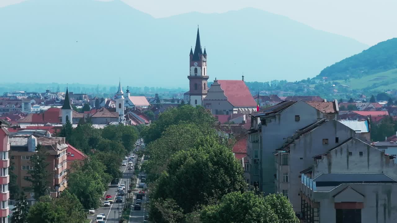 A wide aerial shot of a historical European city center featuring a tall Gothic church tower, surrounded by older buildings, green forested mountains in the background. Bistrita, Evangelical Church