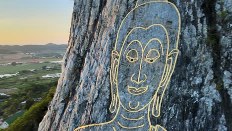 4k close up aerial view: image of buddha, sitting cross-legged, engraved with gold into the northern face of a limestone hill in khao chi chan, thailand