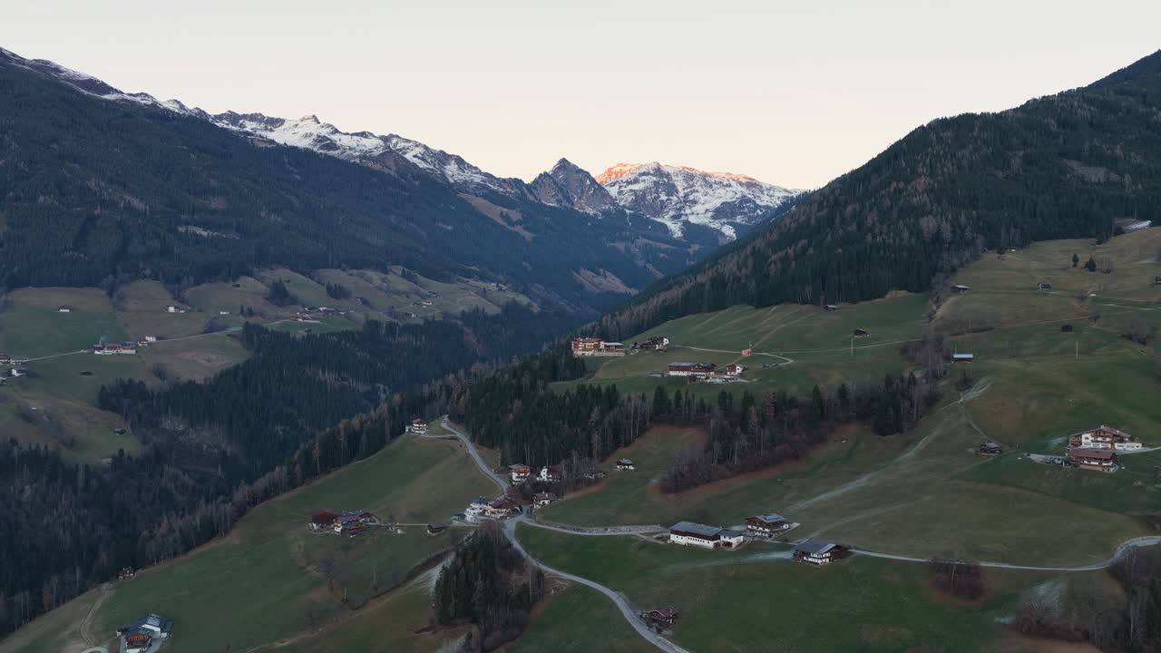 Veduta aerea della valle di Kaltenbach all'alba, con la luce del sole che tocca i pendii e i prati aperti tra le cime alpine. La nebbia si posa bassa sul paesaggio mentre le strutture del villaggio e i sentieri diventano più nitidi.