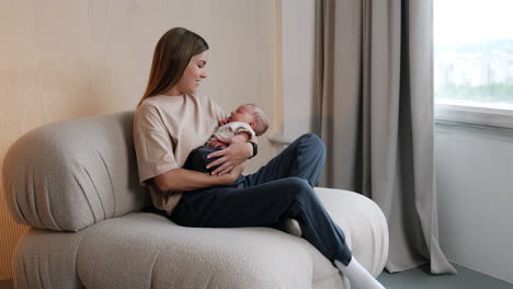 mommy waving her lovely newborn in hands. woman sits on sofa holding her sleeping baby and looking at him with a smile.