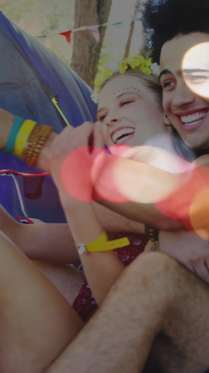 Video verticale: Un uomo si china ridendo, una coppia posa per un selfie a un festival, con cuori che fluttuano intorno.