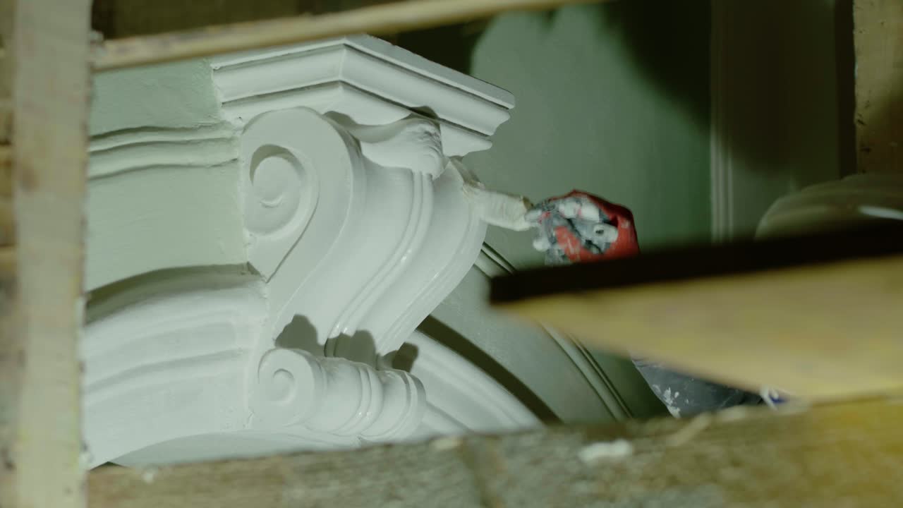 Extreme close-up shot showing a worker's gloved hand carefully painting a detailed, scroll-shaped, white plaster corbel or console bracket during an interior restoration project