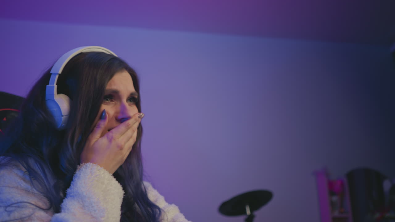 Side view of female live streamer wearing white headset with long dark hair and cozy sweater reacting to negative feedback during stream, seated in dimly lit room with soft purple and blue lighting
