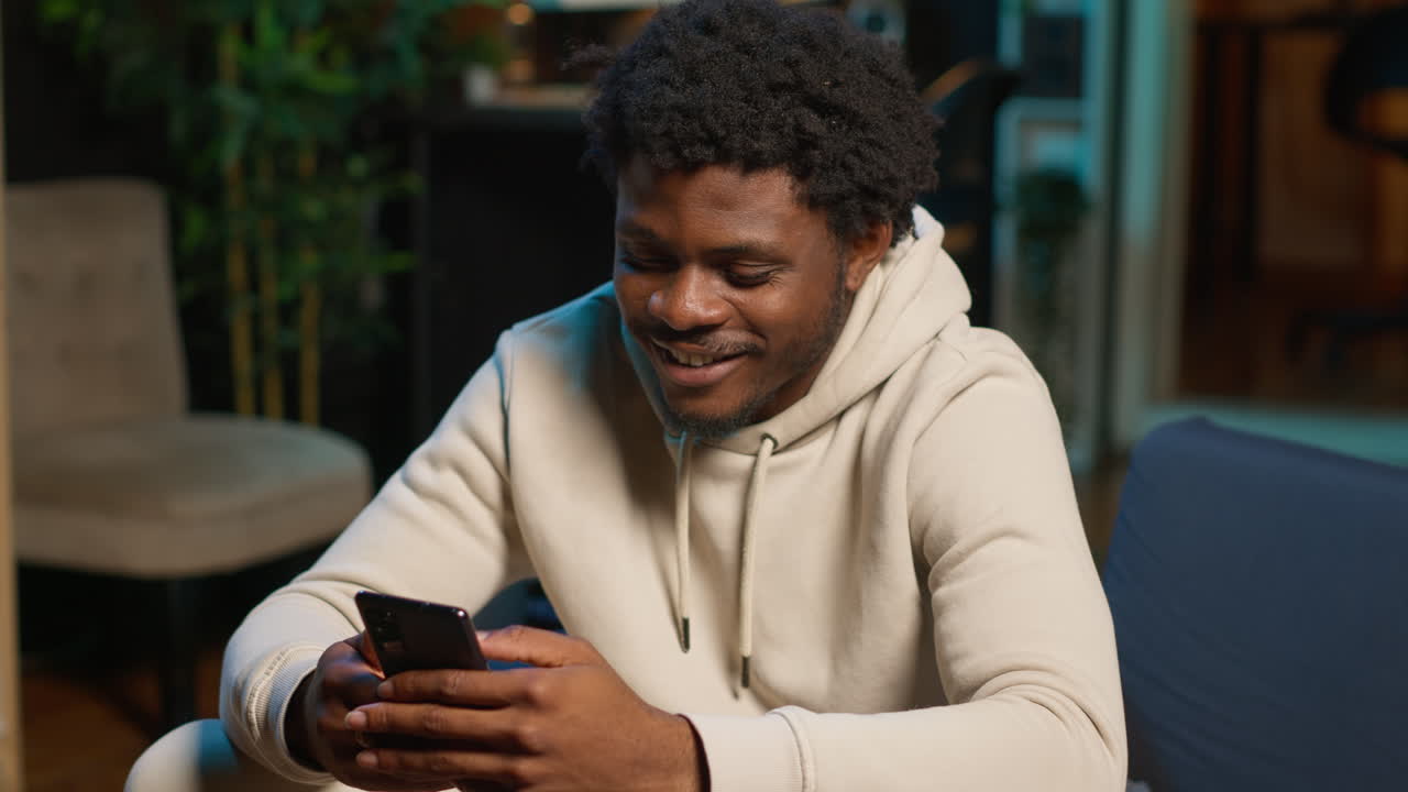 African American man on a sofa texting mates in his living room, entertained by social media and messages. Depiction of remote work breaks, digital communication and relaxation at home. Camera A.