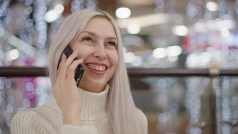 close up fair skinned lady seated on bench holding phone to ear, speaking and laughing heartily with joyful expression, wearing white sweater, jeans and boots against mall interior backdrop