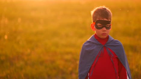 a child in the costume of a superhero in a red cloak runs across the green lawn against the backdrop of a sunset toward the camera.