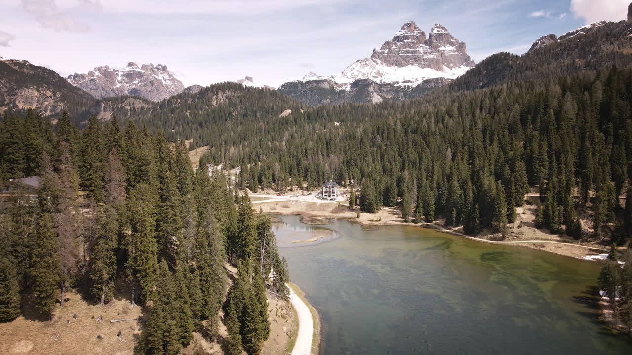 Camera moves forward above a calm alpine lake, passing evergreen forests toward snow‑capped Dolomite peaks and lakeside lodges on a bright spring day
