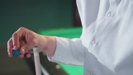 close-up shot of hand in white shirt applying chalk to cue stick before a billiards game. green table and blurred background add depth to focused preparation scene in a dimly lit indoor setting