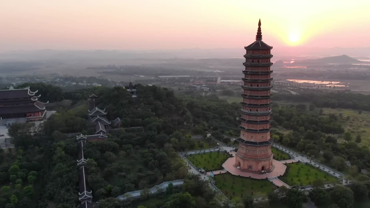 Drone view of a tall traditional pagoda at sunset surrounded by green forest, distant rooftops, and soft glowing light across the countryside and horizon.