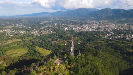 Mobile-Communication-Tower-in-the-mountains,-aerial-view-with-the-city-in-the-background-and-surrounded-with-nature