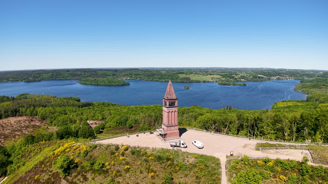 Drone footage captures a historic brick tower standing on a small peninsula by a blue lake in Denmark. The tower and surrounding walkway contrast with lush greenery and calm water