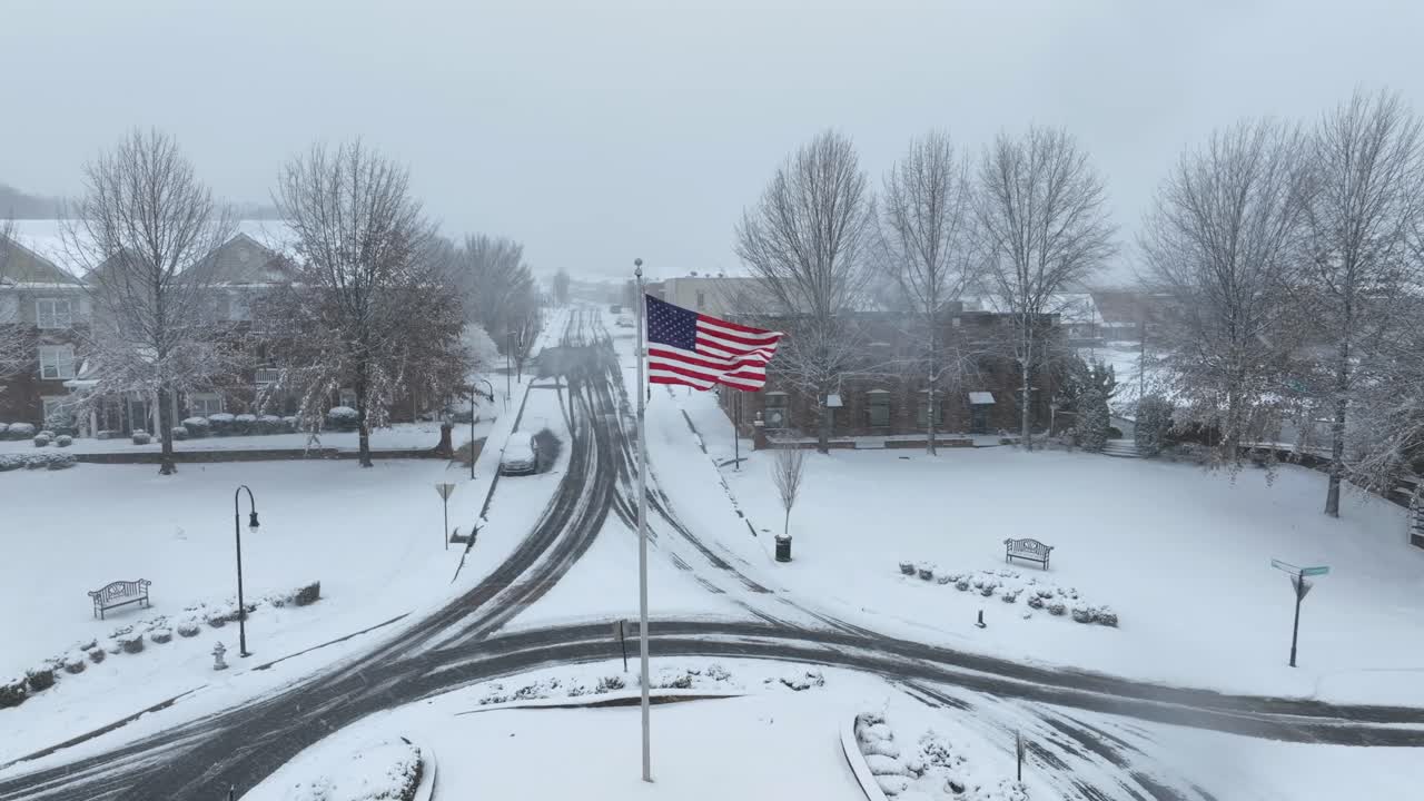 American flag on flagpole in center of snowy roundabout. Aerial approaching shot. Snowy winter snowfall in american town. Empty roads and apartment houses in background