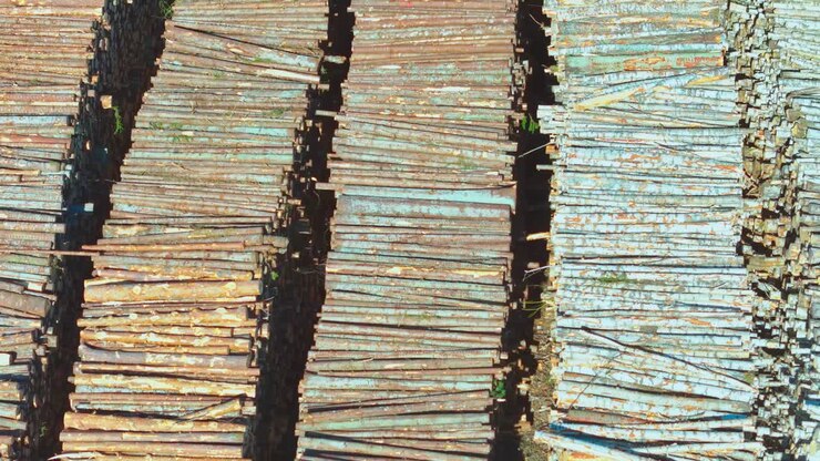 Tumpukan balok kayu lapuk yang tertata rapi tersusun dalam barisan panjang dan sejajar di tempat penggergajian kayu, diambil dari atas dengan variasi warna dan tekstur industrial