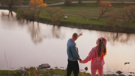 young man and woman looking at the setting sun. romantic couple holding hands while standing on the hill over the river at sunset. rear view.