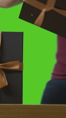 vertical video of man picking up present in gift wrapped box decorated with ribbon on table shot against green screen 1