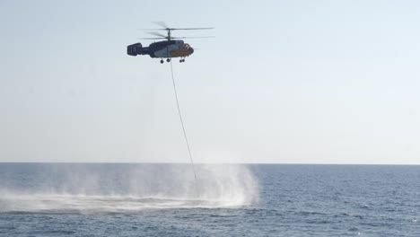 a helicopter with a fire fighting water bucket approaches the water and submerges the bucket to fill with water